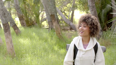 A-young-biracial-woman-with-curly-hair-smiles-in-a-lush-park-setting-with-copy-space