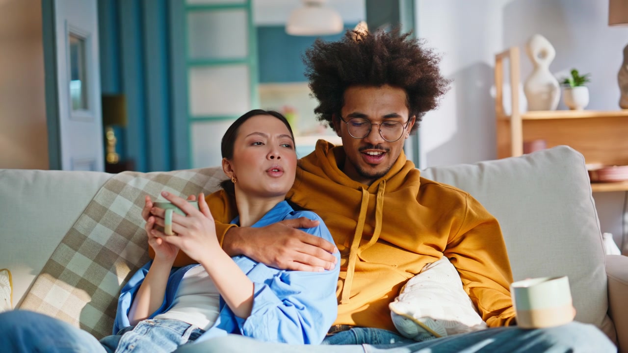 Coppia multirazziale che si rilassa sul divano bevendo caffè e guardando la TV insieme a casa