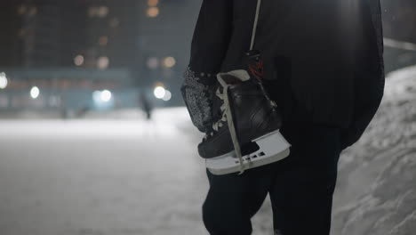 back view of boy walking through snowy outdoor area holding ice skate by lace in hand, winter night atmosphere illuminated by soft glowing city lights and background of skaters in distance