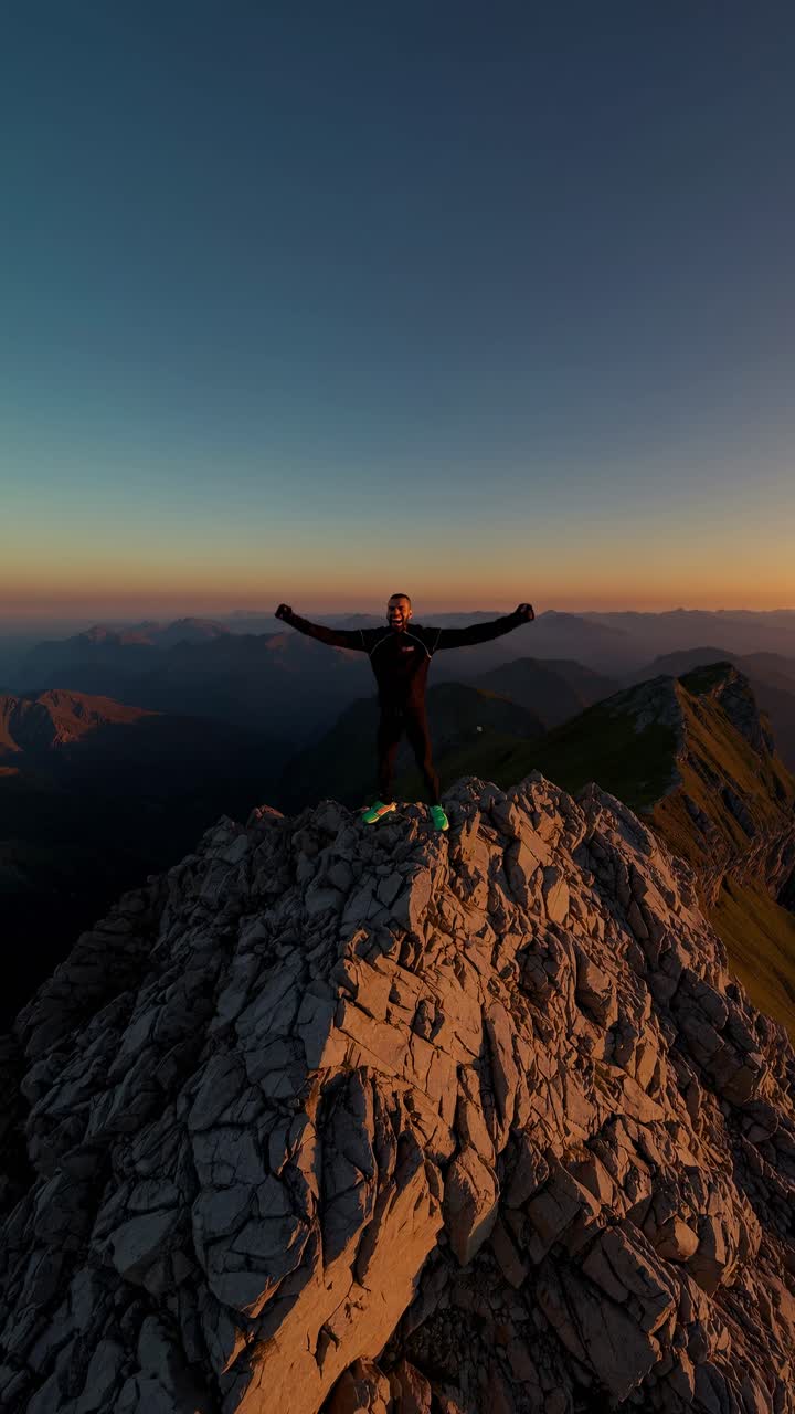 Aerial video shot of a person standing triumphantly on a rocky mountain peak at sunset