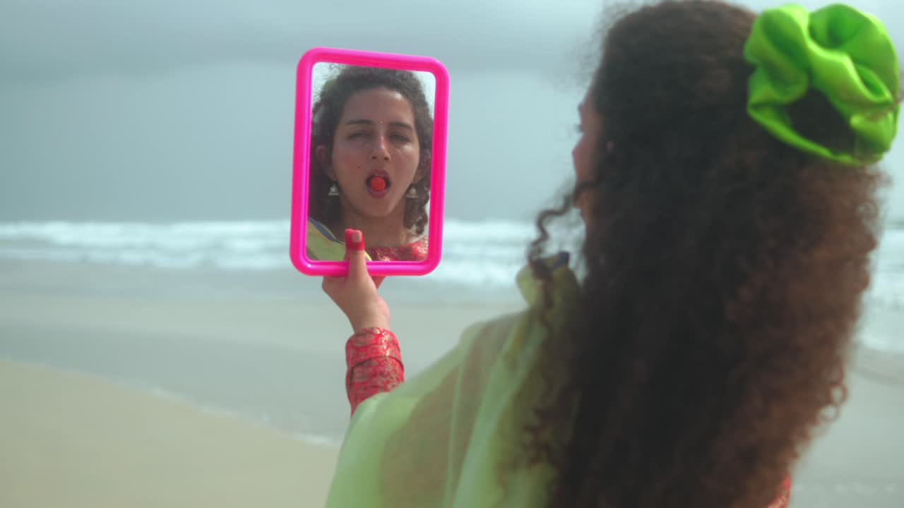 Una donna vista di spalle tiene uno specchio rosa che riflette il suo viso con un frutto rosso in bocca. È in piedi sulla spiaggia con il mare sullo sfondo.