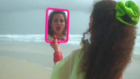 a woman seen from behind holds a pink mirror reflecting her face with a red fruit in her mouth. she's standing on the beach with the sea in the background