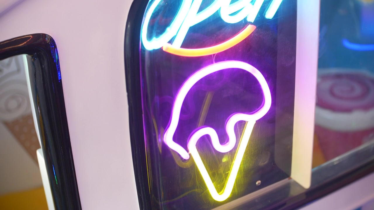Insegna al neon che brilla di blu, rosa e giallo, mostra un messaggio "aperto" con una grafica di un cono gelato. Una vista ravvicinata evidenzia riflessi colorati all'interno di una luminosa gelateria.