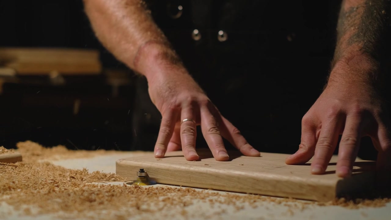 Slow motion of craftsman guiding wooden board over router bit, shaping timber edge as wood shavings scatter across workbench during woodworking process