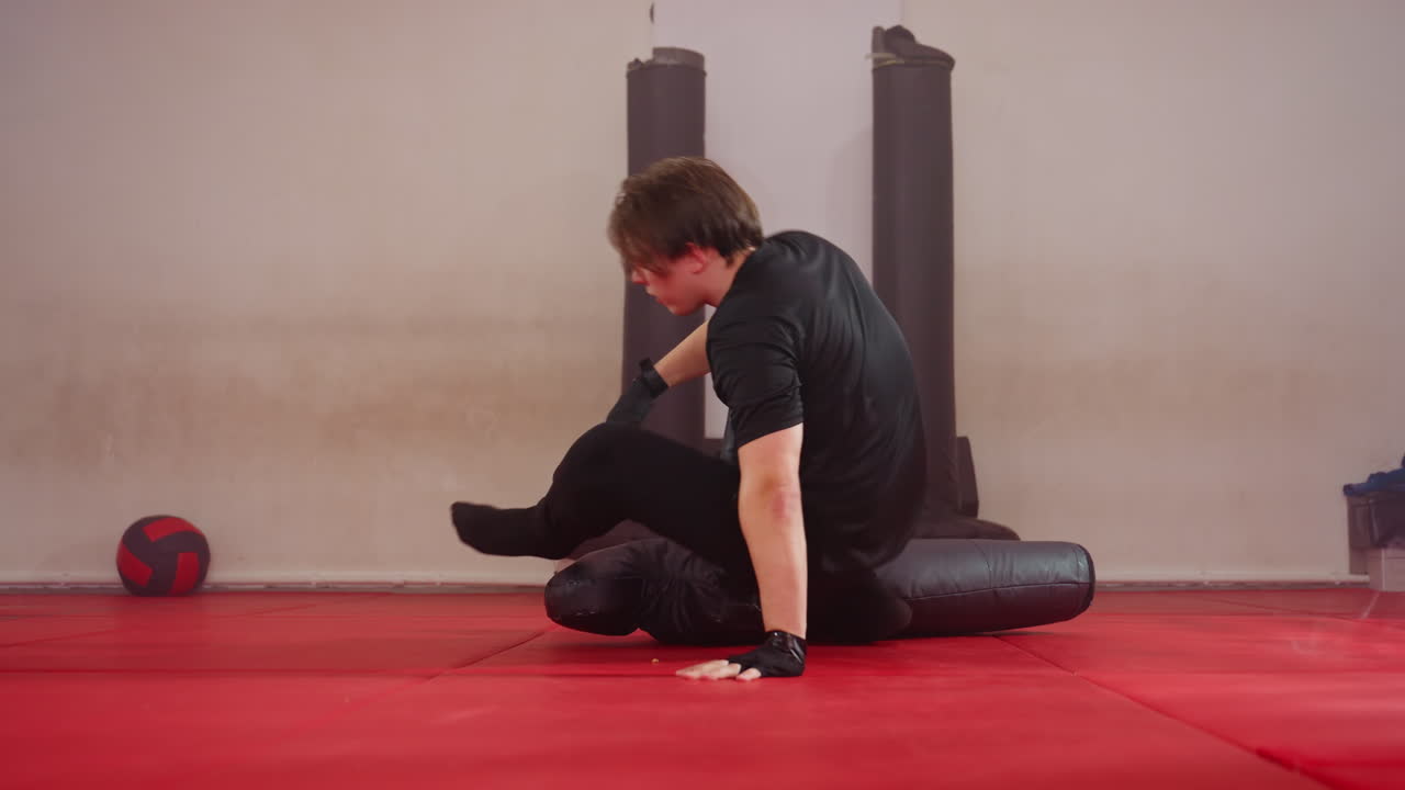 Male fighter practicing ground combat techniques in gym, wearing black sportswear, training with dummy on red mat, performing grappling moves with focus building strength, endurance, martial arts discipline