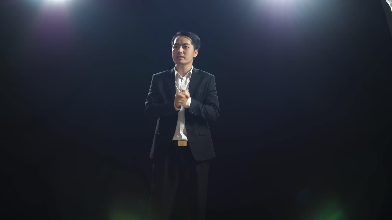 un orador asiático en traje de negocios sosteniendo sus manos juntas mientras habla en el estudio de pantalla negra