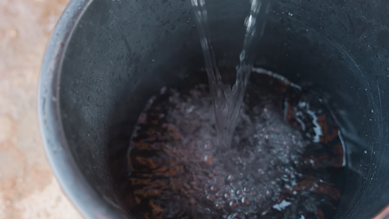 Secchio nero che si riempie d'acqua limpida, con il fondo arrugginito visibile e un getto costante che schizza all'interno. Preparazione dell'officina per la pulizia di falegnameria, risciacquo di pennelli e attrezzi. Pavimento in cemento esterno, texture ravvicinata.