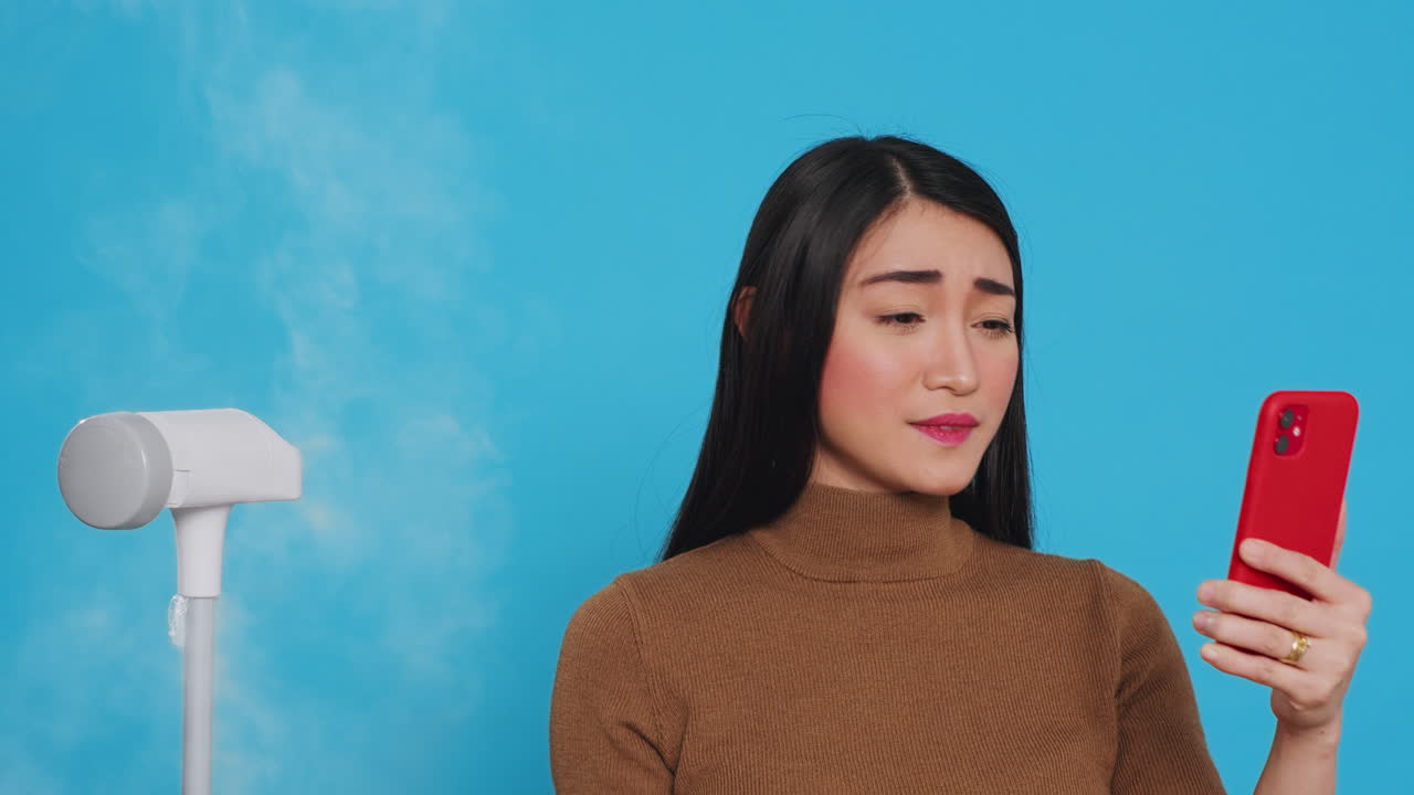 Professional housekeeper ironing clothes using steamer while having videocall meeting on phone,