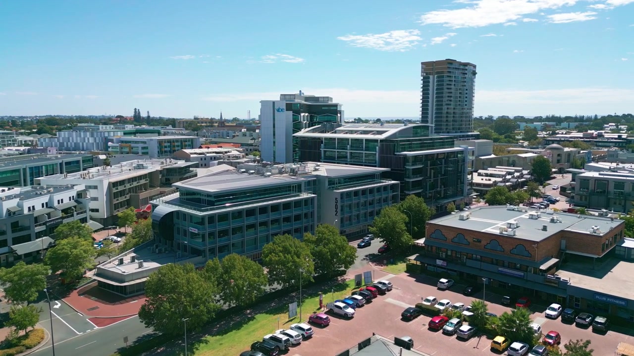 Volo in avanti sopra il quartiere commerciale di Subiaco con moderni edifici per uffici, negozi al dettaglio, parcheggi e sobborghi residenziali che si estendono fino all'orizzonte sotto un cielo blu. Subiaco, Perth, Australia.
