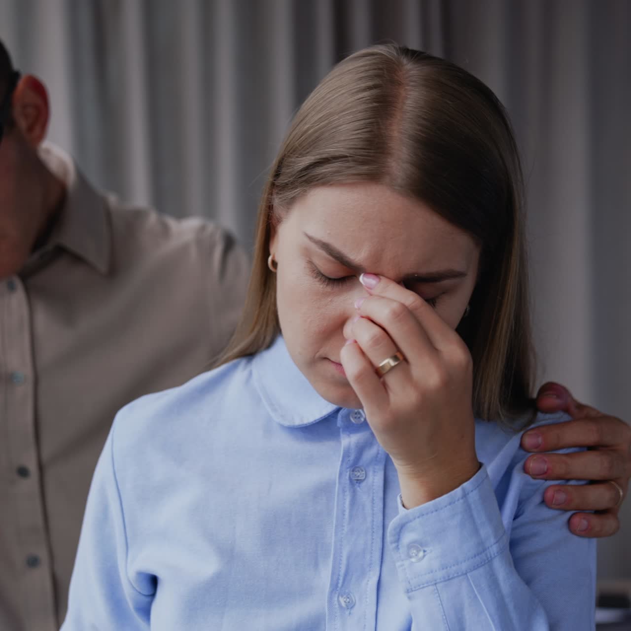 Man is holding a lady by her shoulders from behind. Lady feeling bad and rubs her nose bridge. Male colleague talks and comforts lady