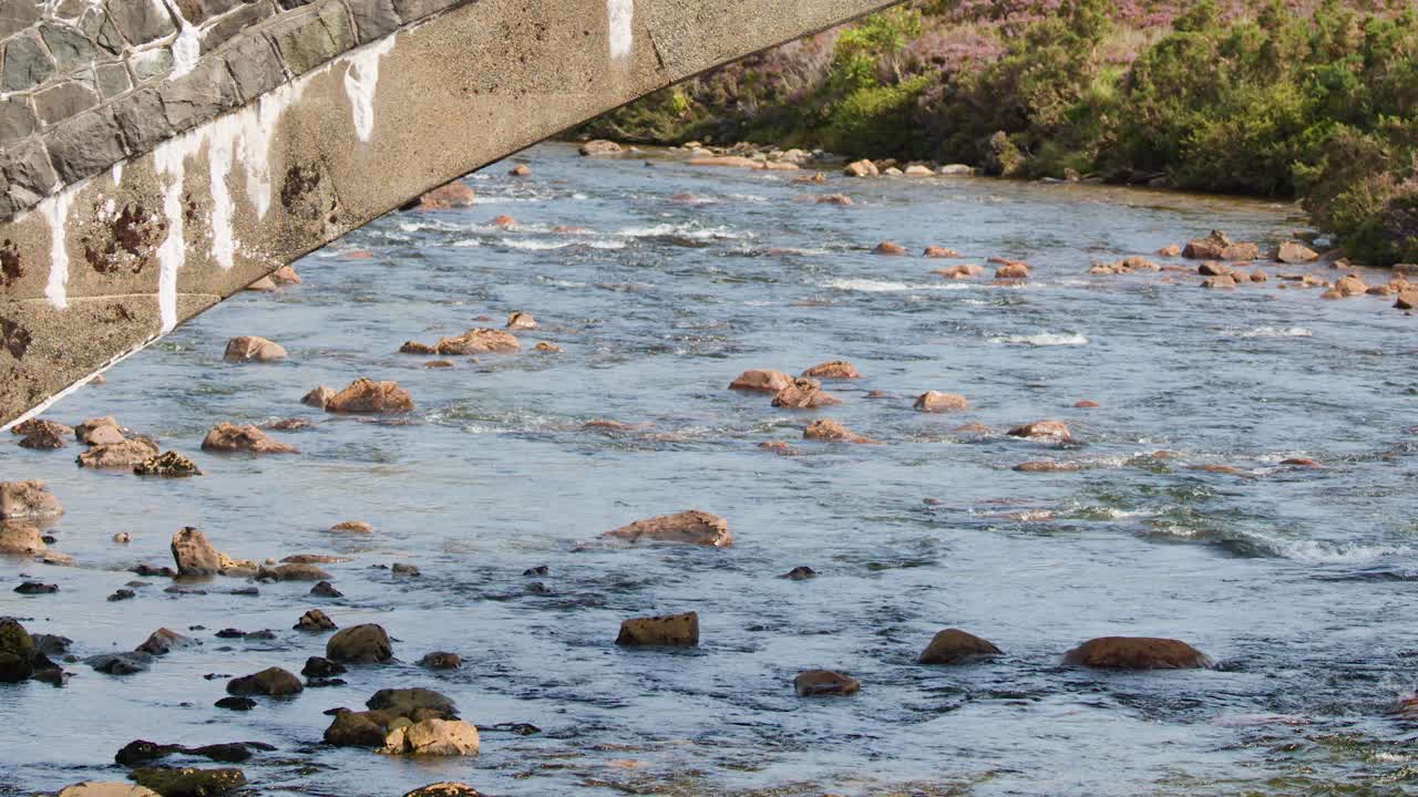 Camera pans across stone bridge and flowing river in bright daylight, Highland landscape visible