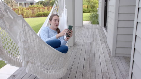 Happy-caucasian-woman-lying-in-hammock-and-using-smartphone-in-garden-in-slow-motion