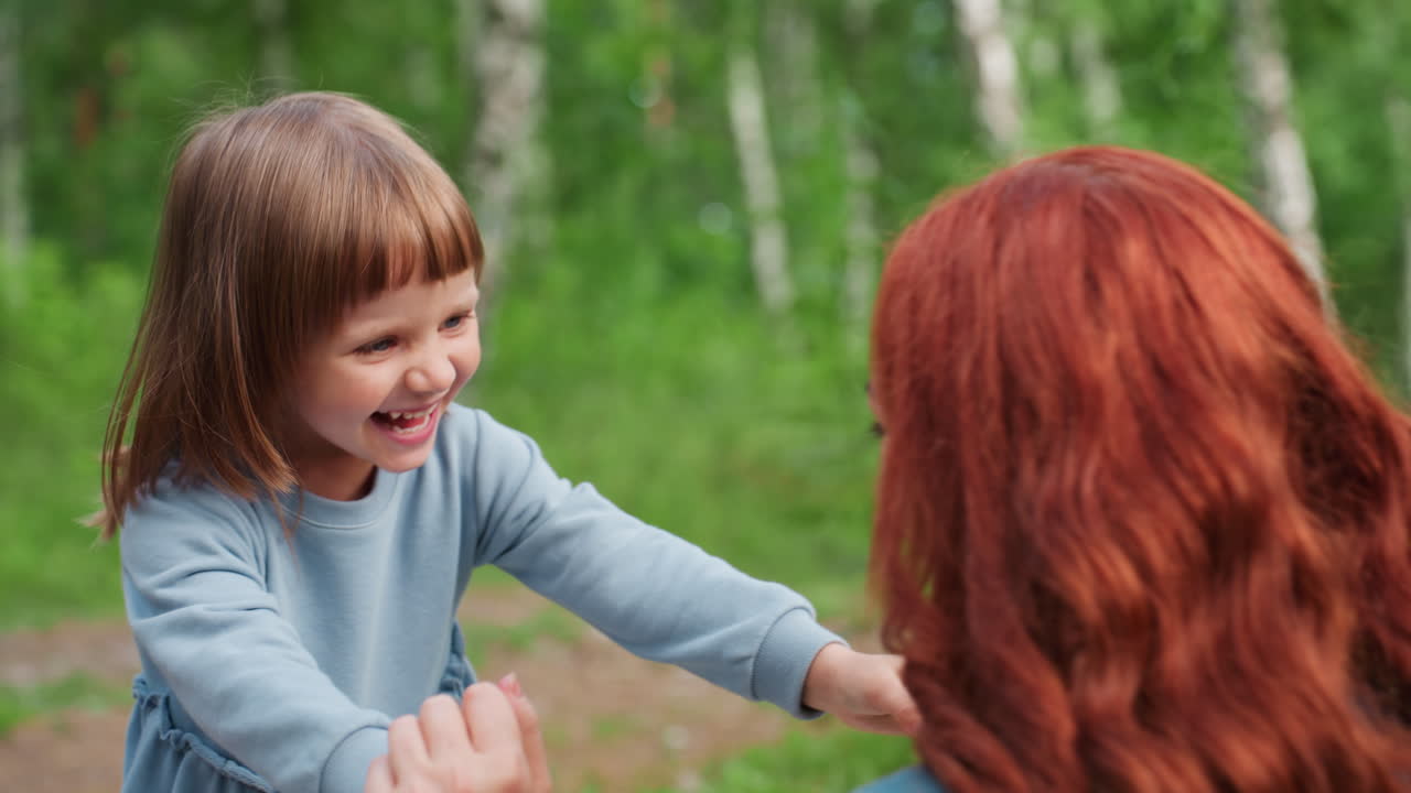 푸르른 숲으로 둘러싸인 야외에서 엄마와 손뼉을 치며 장난치고, 밝게 웃으며 따뜻함, 사랑, 기쁨을 명랑한 웃음으로 표현하는 행복한 어린 소녀