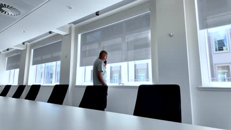 un hombre está de pie junto a la ventana en una sala de conferencias moderna mientras habla en su teléfono, iluminado por la luz natural que se filtra a través de las grandes ventanas, creando un espacio de trabajo sereno
