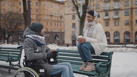 Muslimische-Frau-Und-Ihr-Behinderter-Freund-Im-Rollstuhl-Trinken-Kaffee-Zum-Mitnehmen-Auf-Einer-Bank-In-Der-Stadt-Im-Winter-4