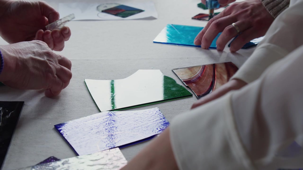 A teacher and her students are working on stained glass projects at a table in the sun. The table is full of colored glass and everyone reaches for tools or materials.