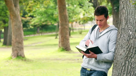 Man-leaning-against-a-tree-as-he-reads-a-book-before-looking-at-the-camera-while-smiling