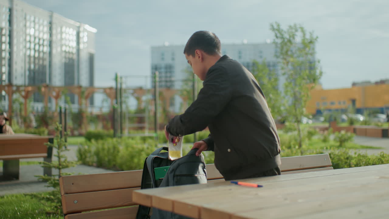 Boy outdoors packing study materials into backpack after finishing study session at wooden bench, open book and pens left on table, blurred person visible in background