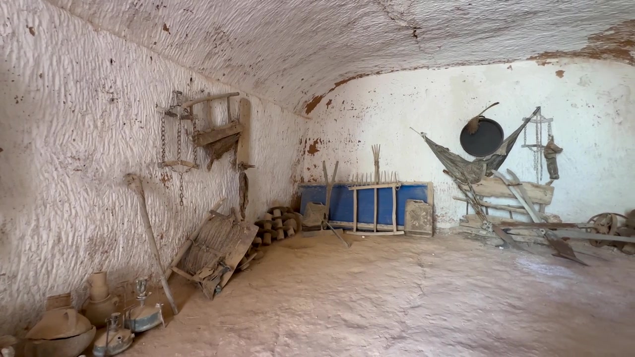 Una stanza sotterranea di Matmata con pareti in argilla ospita attrezzi agricoli rustici, slitte di legno, vasi e un'amaca, evocando la tradizionale vita berbera del deserto e gli interni rurali storici. Tunisia