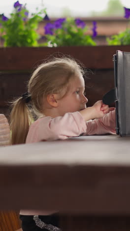 serious blonde little girl surfs internet on contemporary tablet computer sitting at large rustic table in hotel cafe on rainy day slow motion