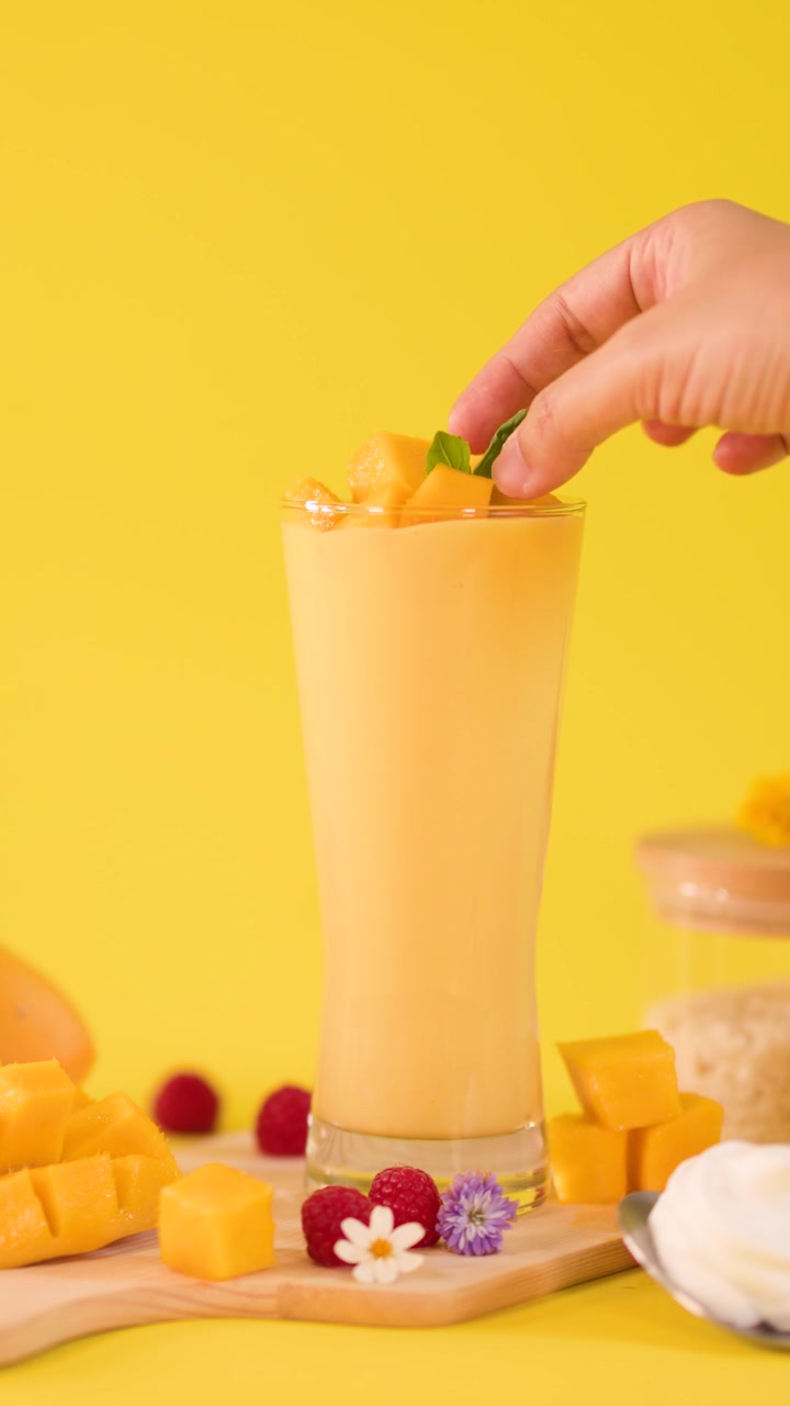 Una mano umana aggiunge una guarnizione di menta a un frullato alto al mango tra frutta tropicale su sfondo giallo.