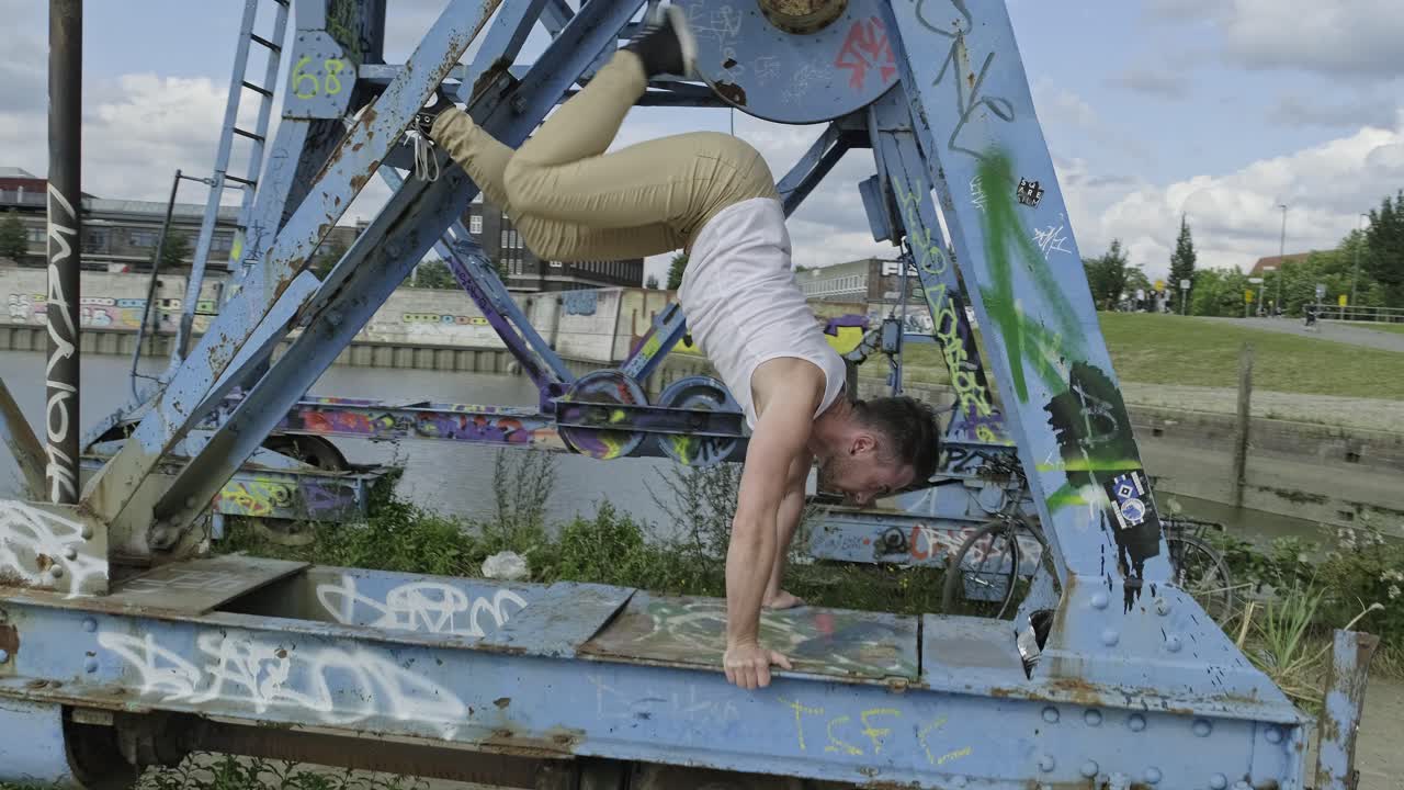 Pan left side view of young hipster guy with sporty figure performing handstand while practicing acrobatic dance movements on weathered metal structure in city
