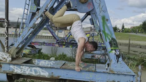 pan left side view of young hipster guy with sporty figure performing handstand while practicing acrobatic dance movements on weathered metal structure in city