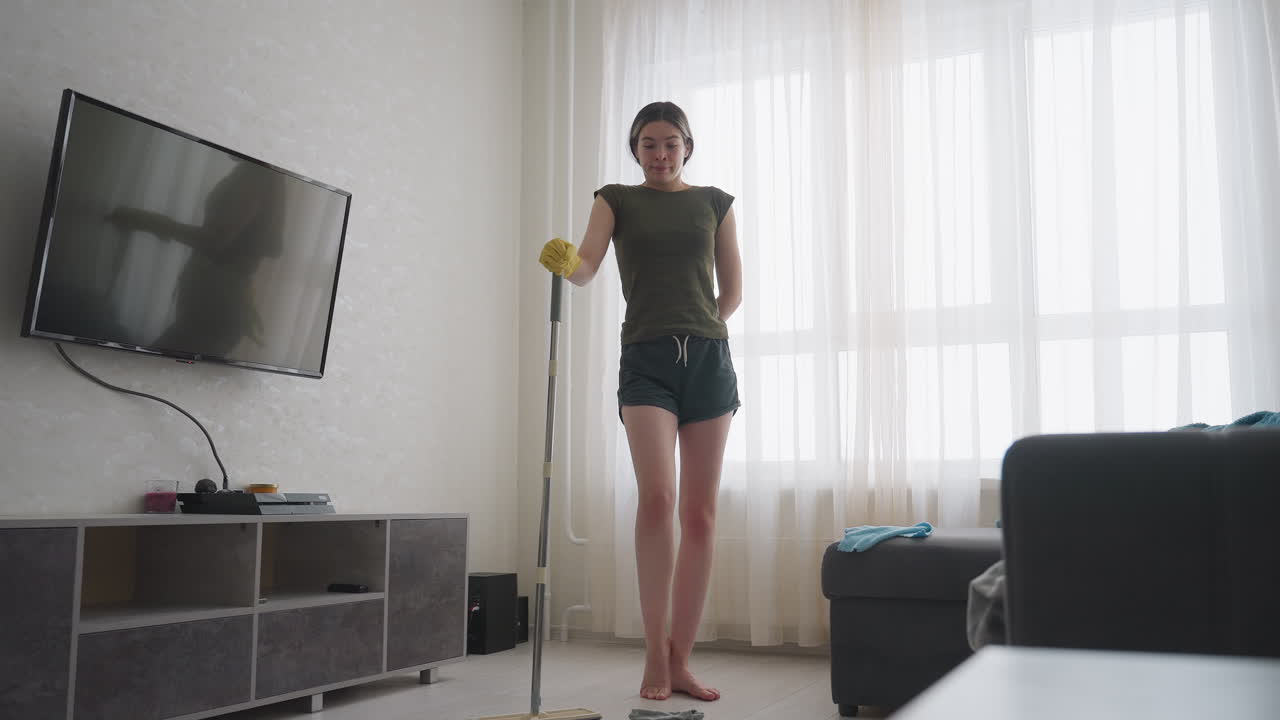 Maid in casual outfit wearing gloves stands with mop watching messy living room, clothes scattered on floor and sofa, her reflection visible on television screen while she shakes head in surprise