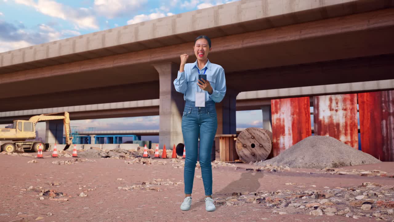 Cuerpo completo de una mujer asiática con su smartphone en una obra de construcción de una autopista. Levanta el puño y grita un gol.