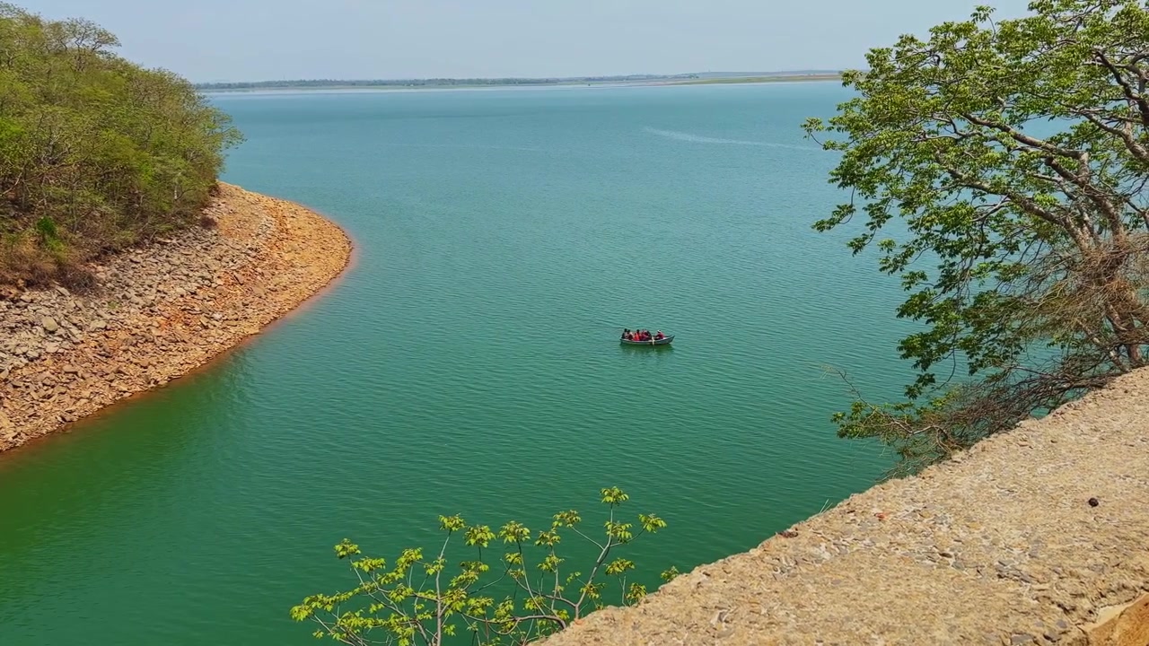 Ampia inquadratura lunga di turisti indiani che si godono un giro in pedalò attraverso un tranquillo bacino idrico blu circondato da una costa rocciosa e una foresta lussureggiante.