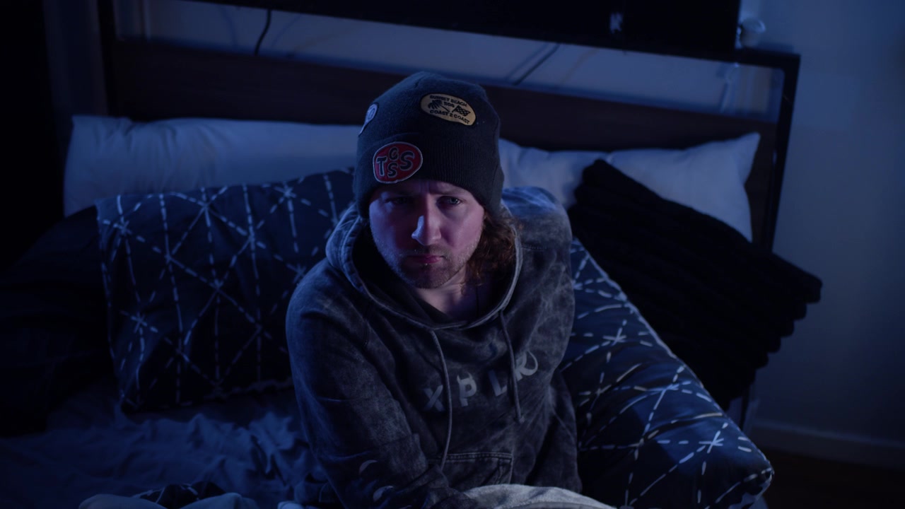 Cena cinematográfica 4K de um homem sentado na cama, olhando para o armário, antes de reagir com medo a algo atrás da câmera, recuando de uma presença invisível em seu quarto iluminado pela lua.