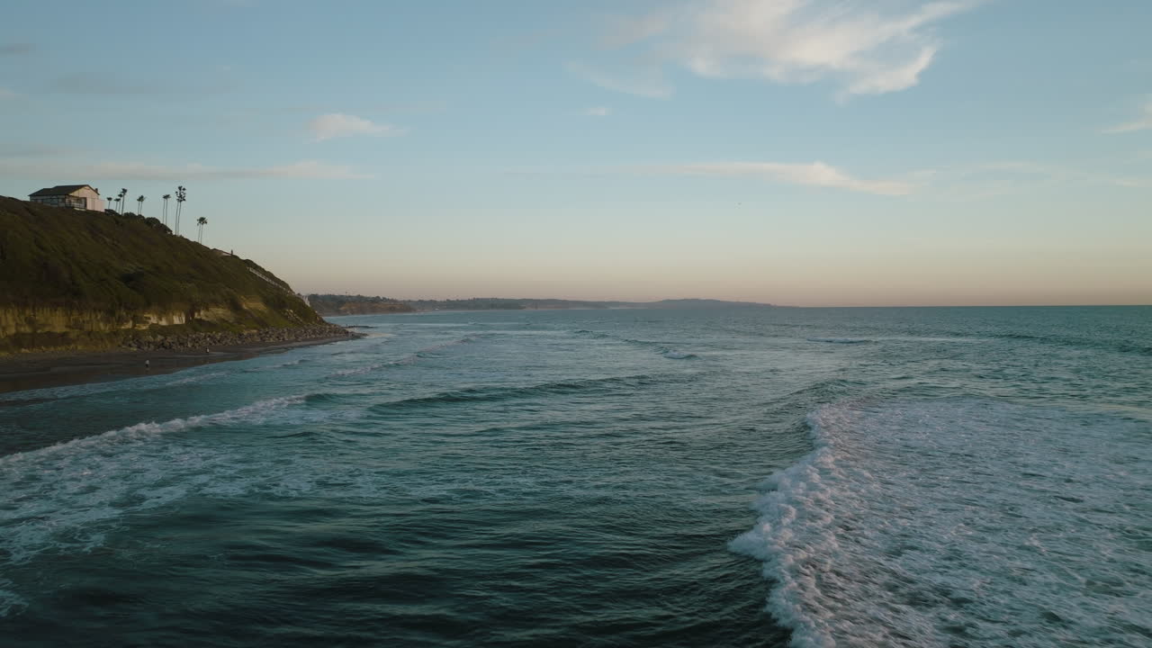 Aerial dolly over ocean waves crashing and crumbling spreading whitewash over California beach with palm trees and homes on cliff