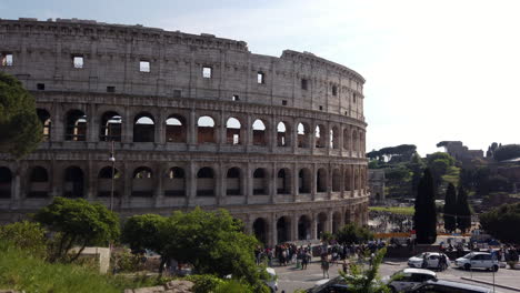 the colosseum from a different side than usual on a sunny spring day. there are few cars parked on the sides of the street. cars are passing by. people are walking on the sidewalk.
