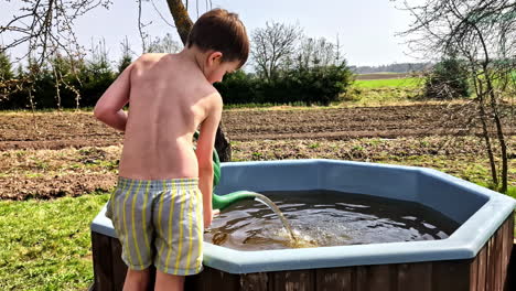 shirtless boy pouring water into outdoor plastic tub from green watering can