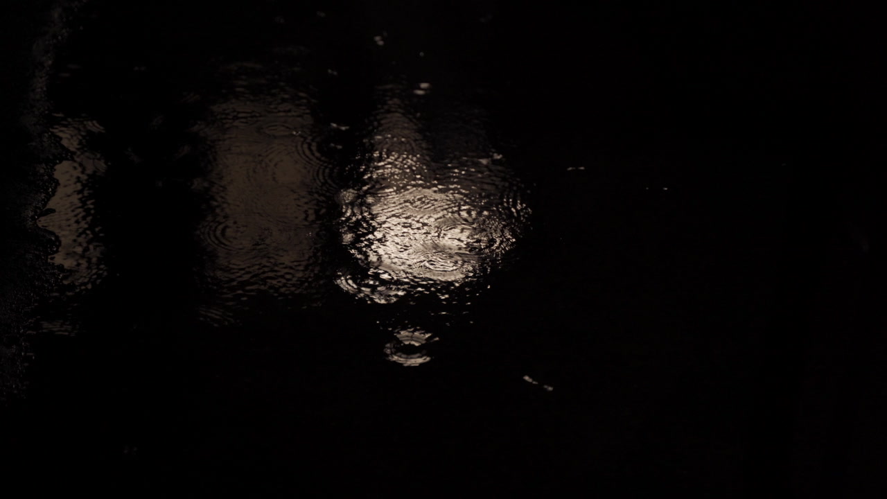 4K cinematic shot of rain falling into a puddle at night with the light of a nearby window reflecting in the rippling water. It could be a storm in any city in the world at night time while raining.