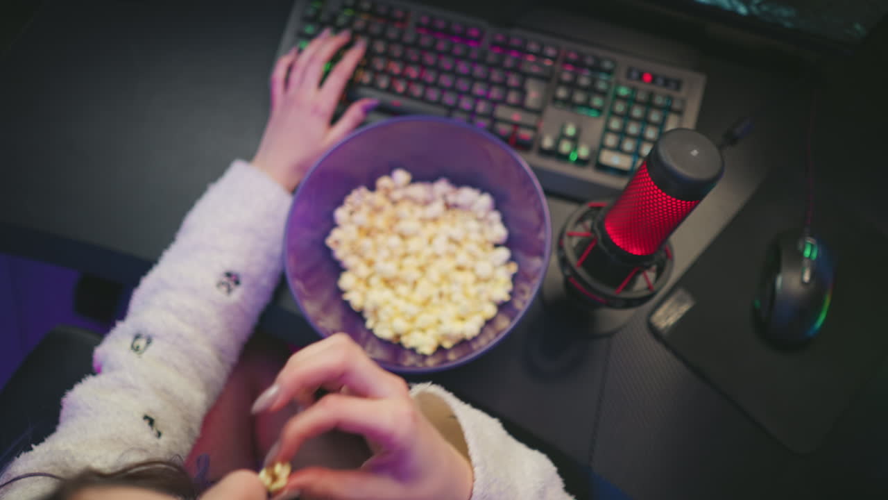 Overhead view of programmer in fluffy sleeves typing and using mouse while eating popcorn from purple bowl, colorful keyboard lighting and red condenser mic visible on desk in vibrant studio setting