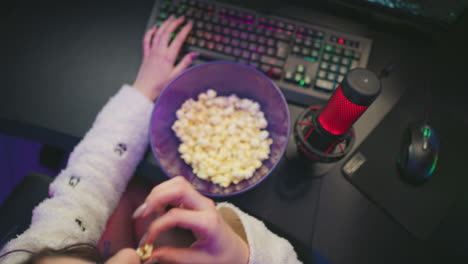 overhead view of programmer in fluffy sleeves typing and using mouse while eating popcorn from purple bowl, colorful keyboard lighting and red condenser mic visible on desk in vibrant studio setting