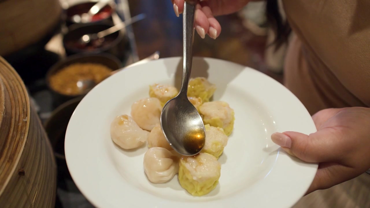 Una persona usa un cucchiaio per versare a filo salsa scura su ravioli al vapore in un piatto.