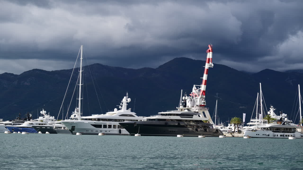 Yacht di lusso bianchi attraccati in un porto con montagne e nuvole temporalesche sullo sfondo. Tivat, Montenegro.