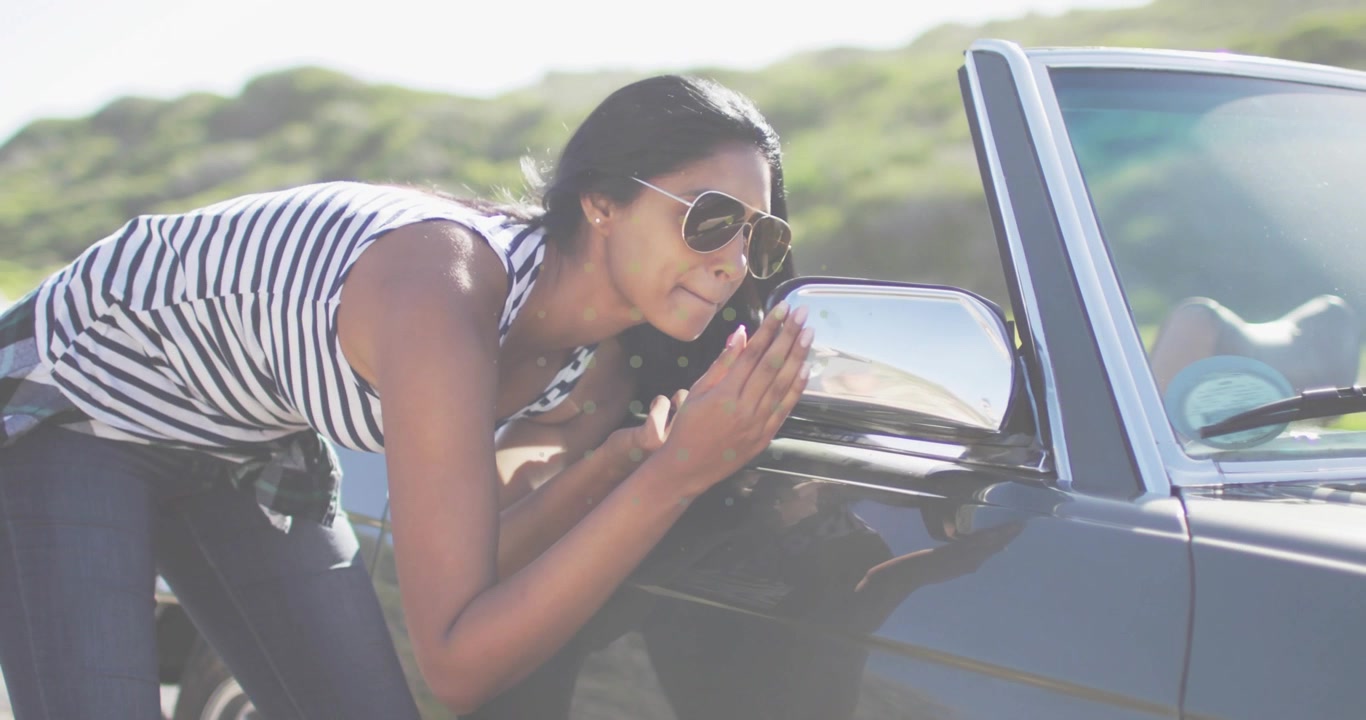 Woman approaching convertible, leaning to driver-side mirror, cupping hands, inspecting reflection