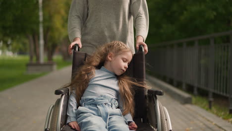 woman pushes wheelchair with little daughter walking in park