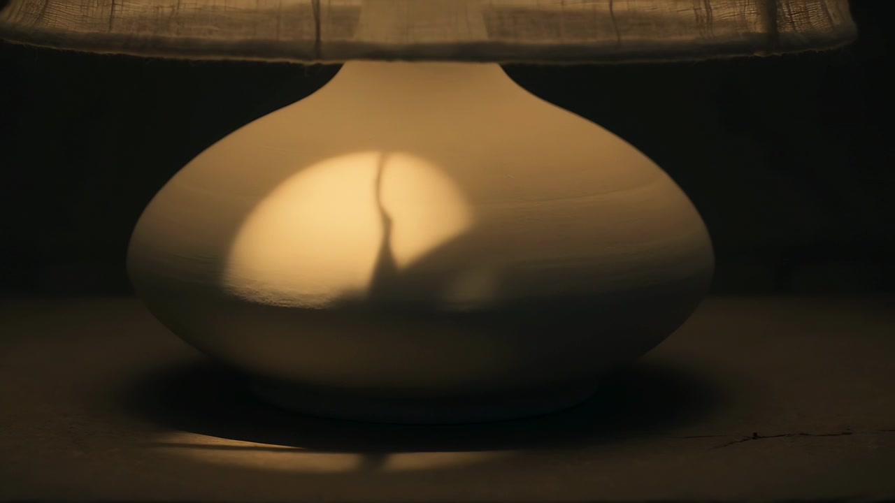 Sich bewegendes, verdeckendes Objekt kreuzt Glühbirnenstrahl und erzeugt geschichtete Schatten auf Keramiklampe auf Tischplatte.