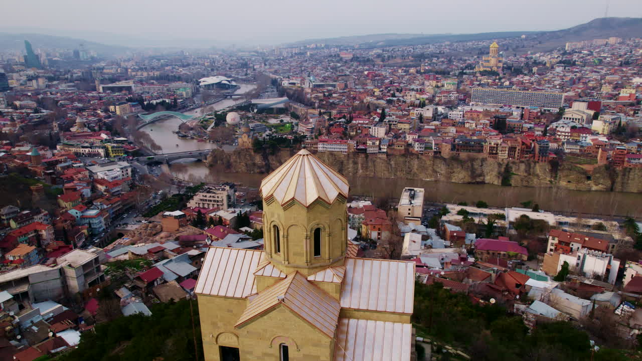 Plynulý let dronu nad kopulí kostela, nad Starým Městem Tbilisi v Gruzii, letecký pohled na panorama města a řeky zdůrazňuje náboženské dědictví, hustotu zástavby a dramatický terén střech podél řeky Kury.