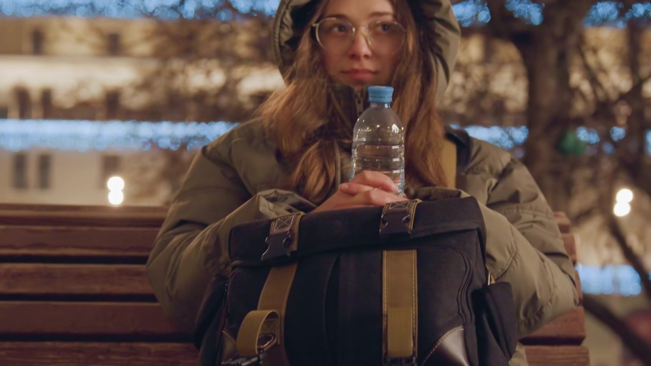 Girl resting outdoors. Caucasian girl enjoying winter moment. Young woman in park during peaceful snowy evening. Sitting on bench with water bottle amidst snowy winter evening lights