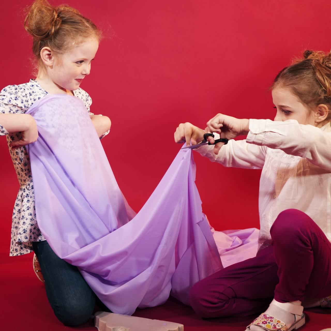 two little girls are playing with a lilac cloth on a pink studio background. One of girl is cutting fabric by scissors in her hand