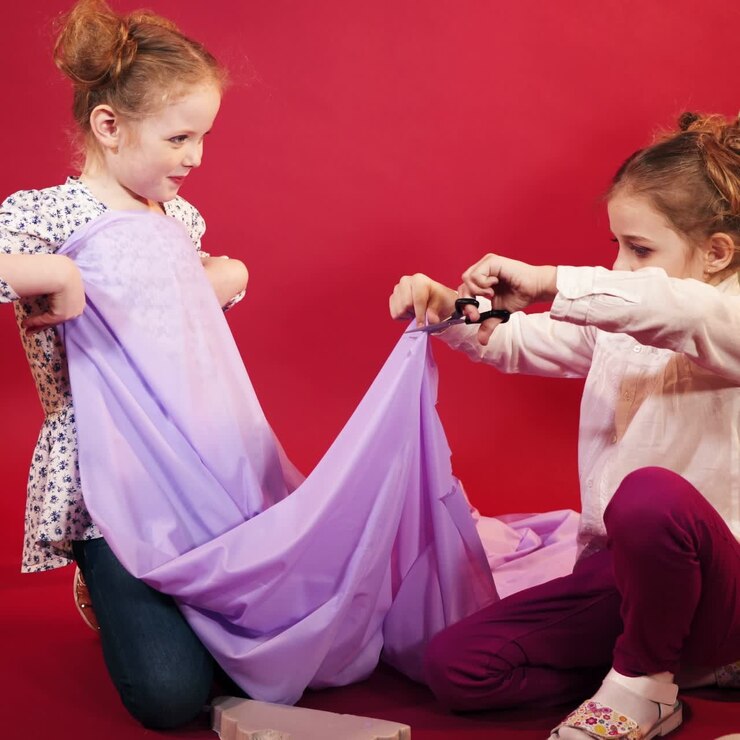 two little girls are playing with a lilac cloth on a pink studio background. One of girl is cutting fabric by scissors in her hand