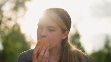 lady wearing grey top enjoys eating burger with a thoughtful expression in sunlit outdoor setting, soft light illuminating her face, background is blurred with warm sunrays