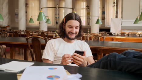 Junger-Asiatischer-Männlicher-Student,-Der-Mobiltelefon-In-Der-Bibliothek-Benutzt