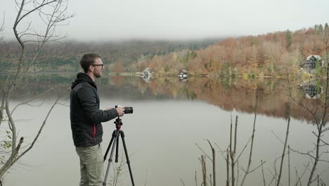 Hombre-Organizando-Su-Cámara-Para-Capturar-Una-Imagen-De-Un-Hermoso-Lago-Reflejado-Y-Colores-Otoñales-Perfectos-En-El-Fondo-Y-Niebla-Baja-En-El-Fondo