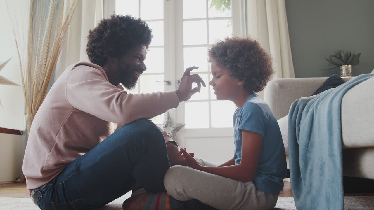 Close up of a middle aged black father and his pre teen son sitting cross legged on their living room floor opposite each other, playing games and laughing, close up